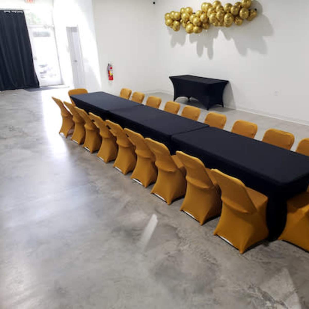A long table covered with a black cloth is surrounded by yellow chairs in a spacious, well-lit room.