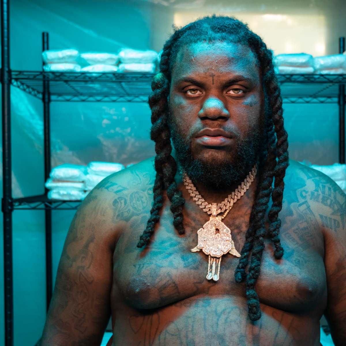 A shirtless man with long braids stands in front of stacked bags on shelves within a brightly lit room.