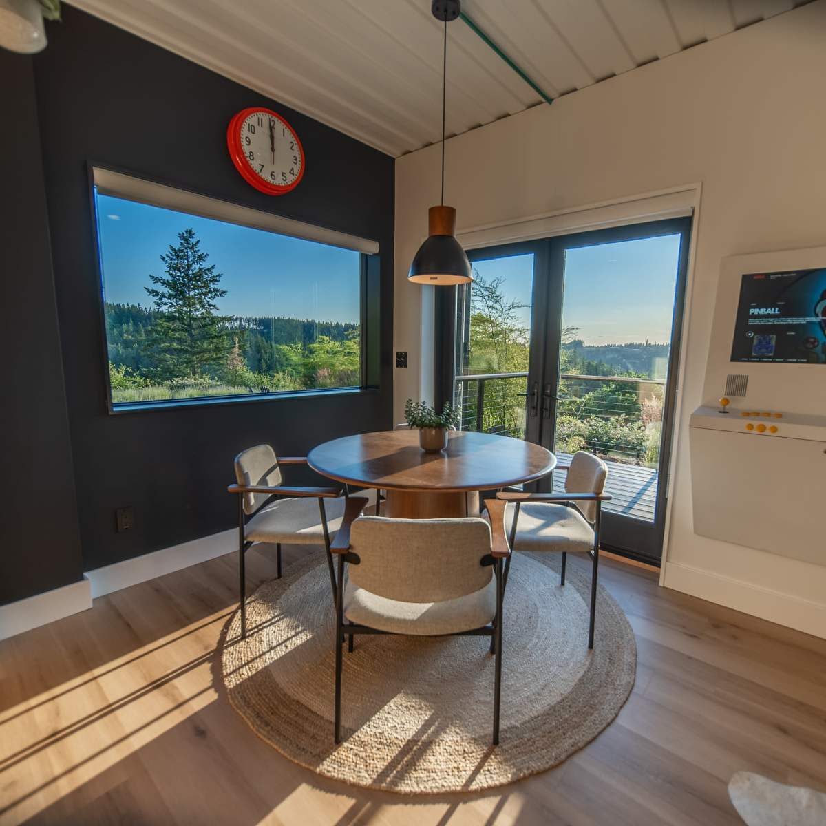 A modern dining area features a circular table with four chairs, large windows showcasing a scenic view, and a wall-mounted TV beside a cheerful clock.
