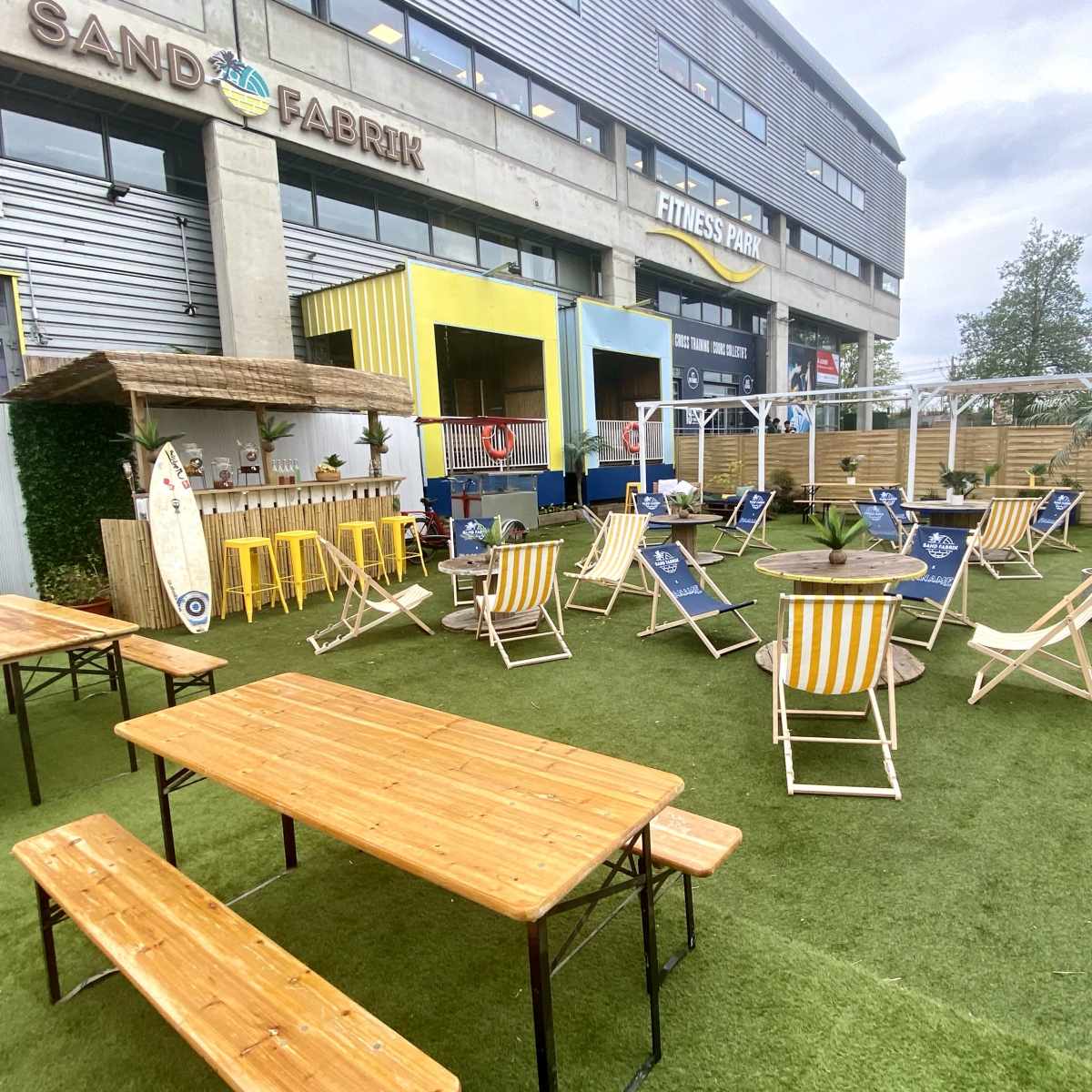 An outdoor seating area features wooden picnic tables, lounge chairs, and greenery, set against a backdrop of a modern building.