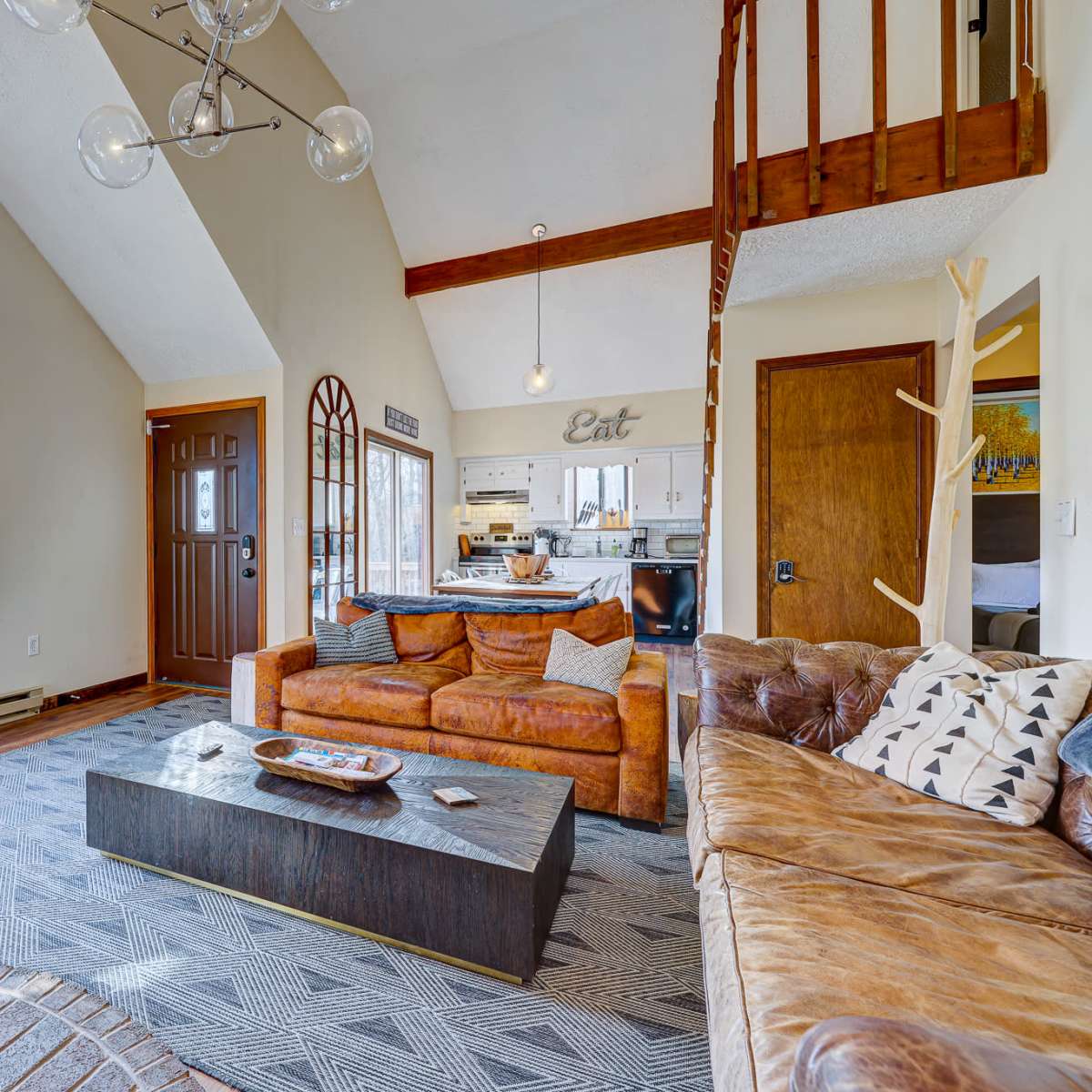 The living room features a combination of a brown leather sofa and an armchair, a coffee table in the center, large windows offering a view of the outdoors, and a stove in the corner.
