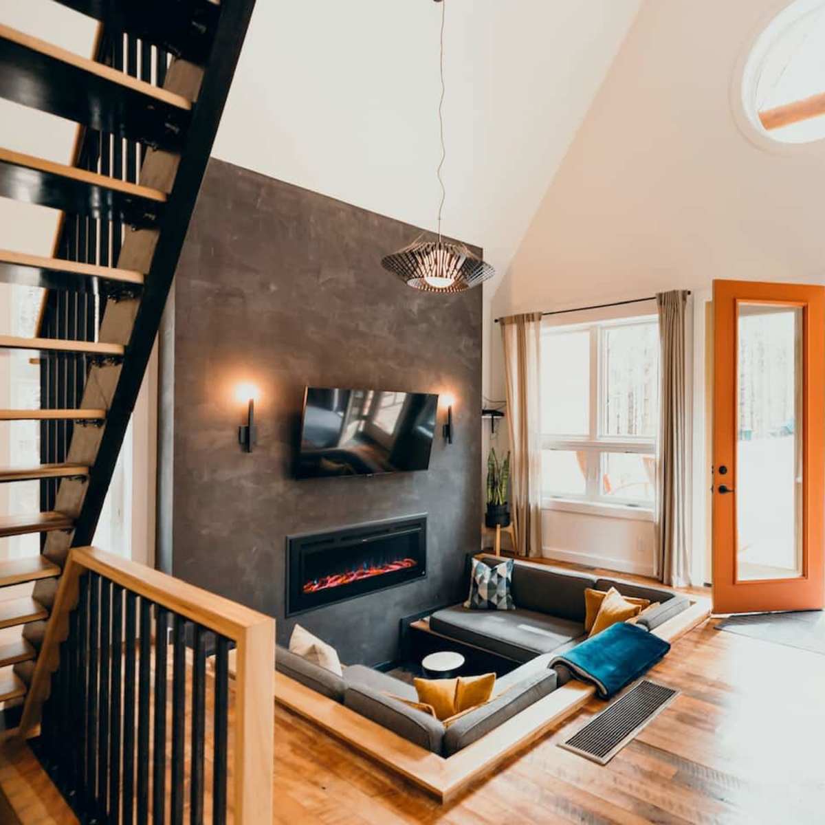 A modern living room with a fireplace, a large wall-mounted TV, and a view of a patio area through orange double doors.