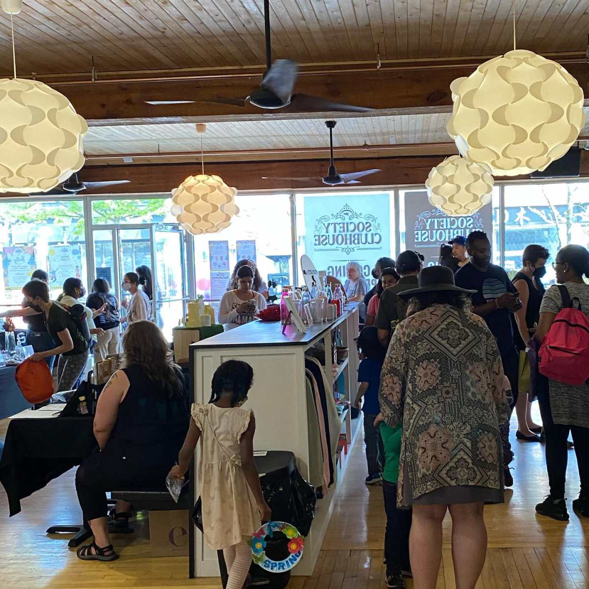 A busy retail space filled with customers browsing products, with large windows and hanging light fixtures overhead.