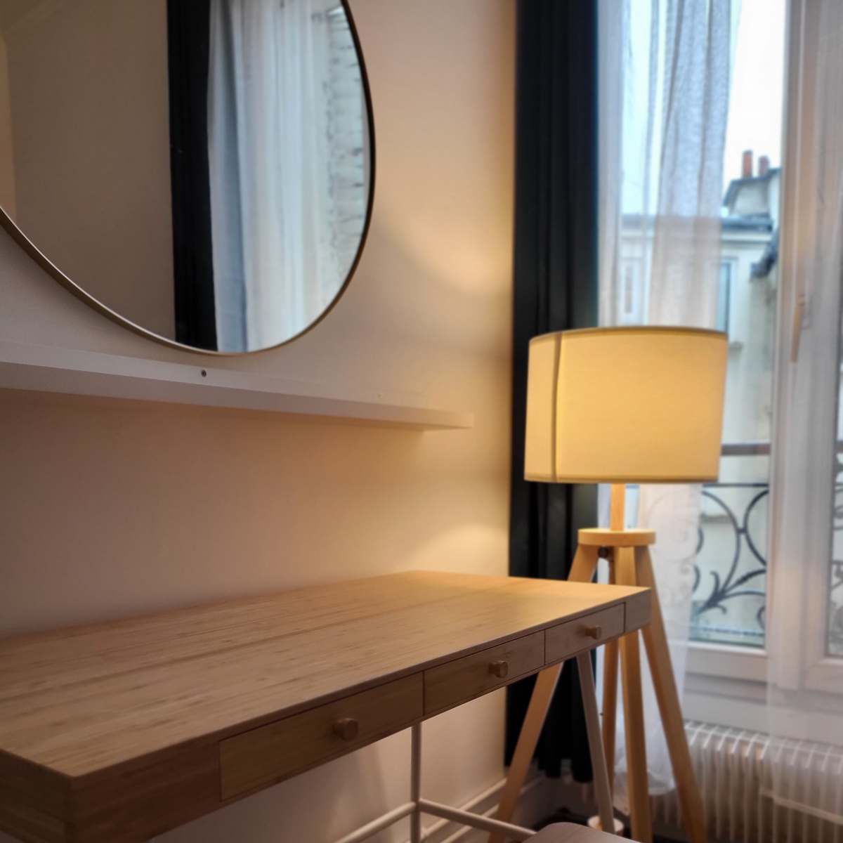 A minimalist workspace featuring a wooden desk and stool with a round mirror above, next to a tall lamp, all set by a window with sheer curtains.