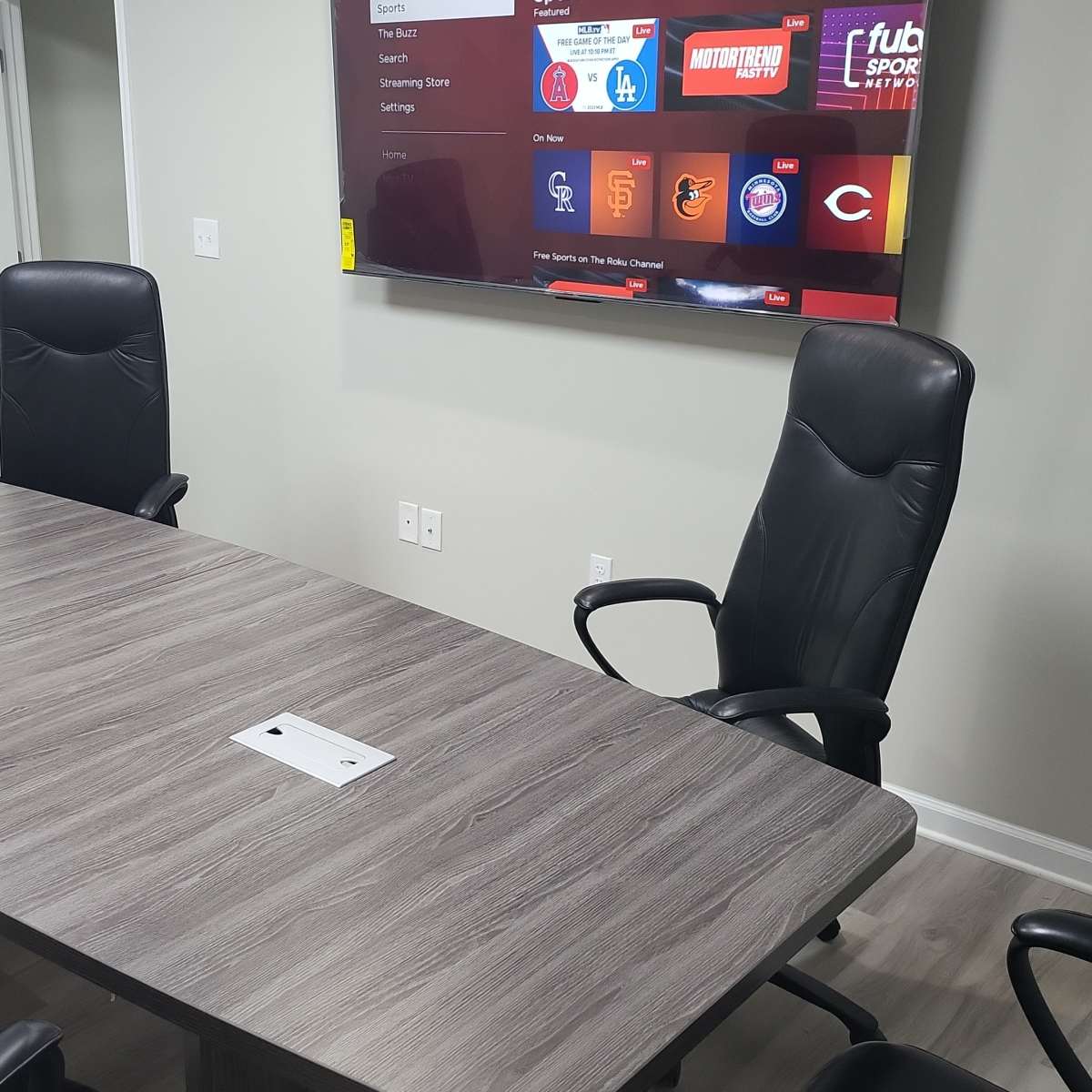 A conference room features a long wooden table surrounded by four black chairs, with a television mounted on the wall displaying various streaming options.
