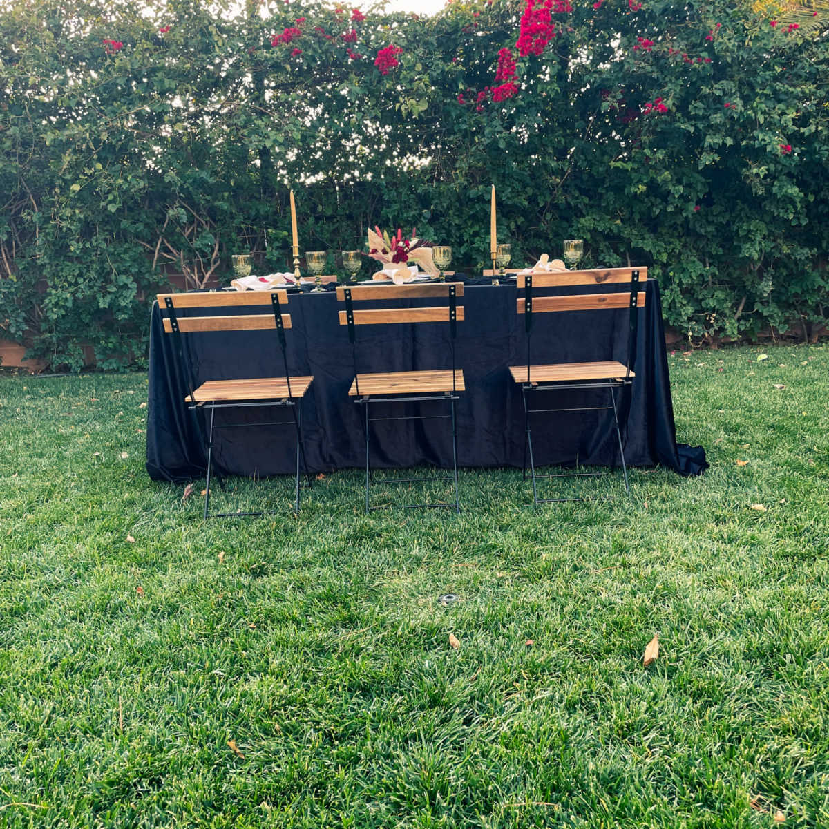 A rectangular table with black tablecloth is set for a meal outdoors, surrounded by four chairs and framed by flowering shrubs.