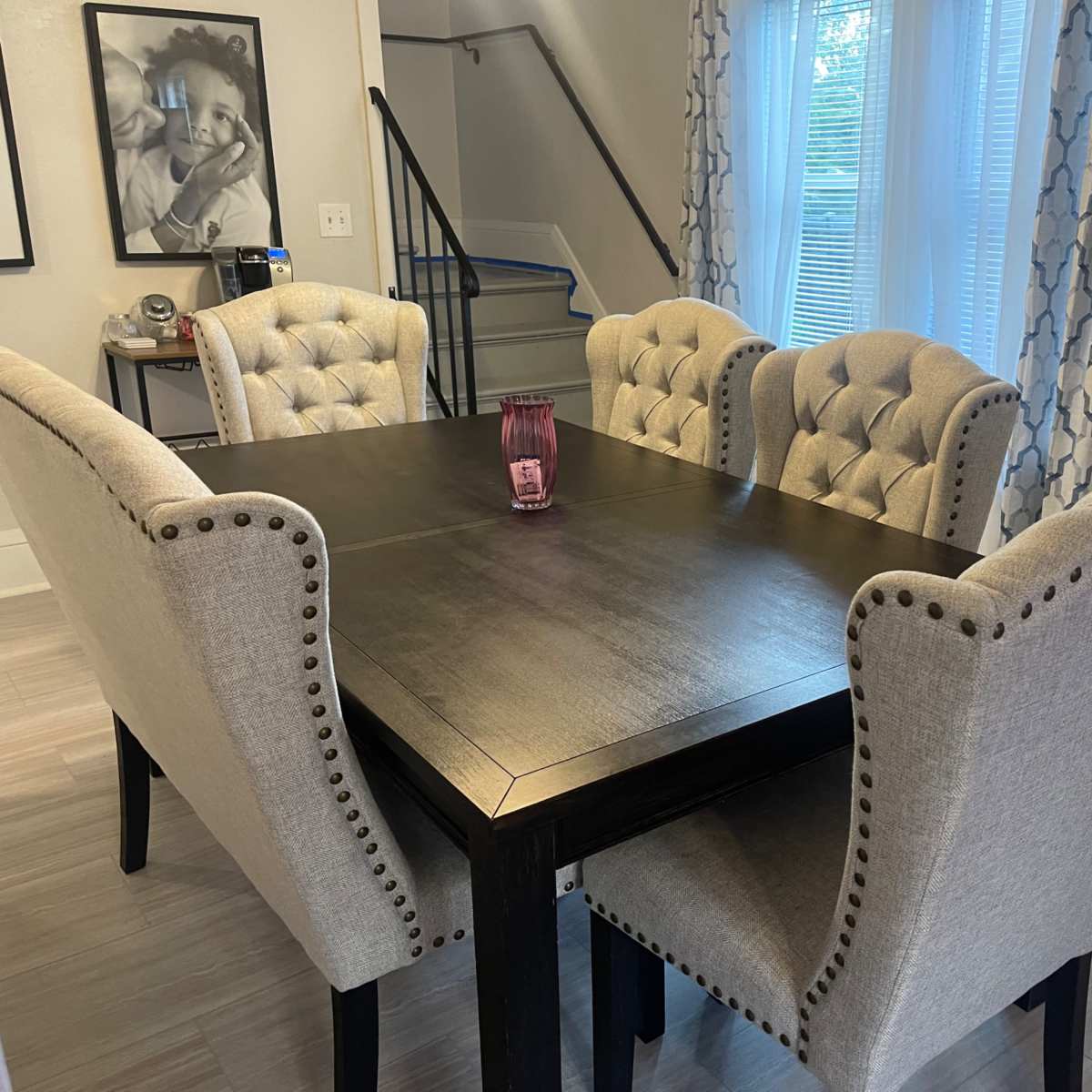 A dining table surrounded by four upholstered chairs sits in a well-lit room with a staircase and window in the background.