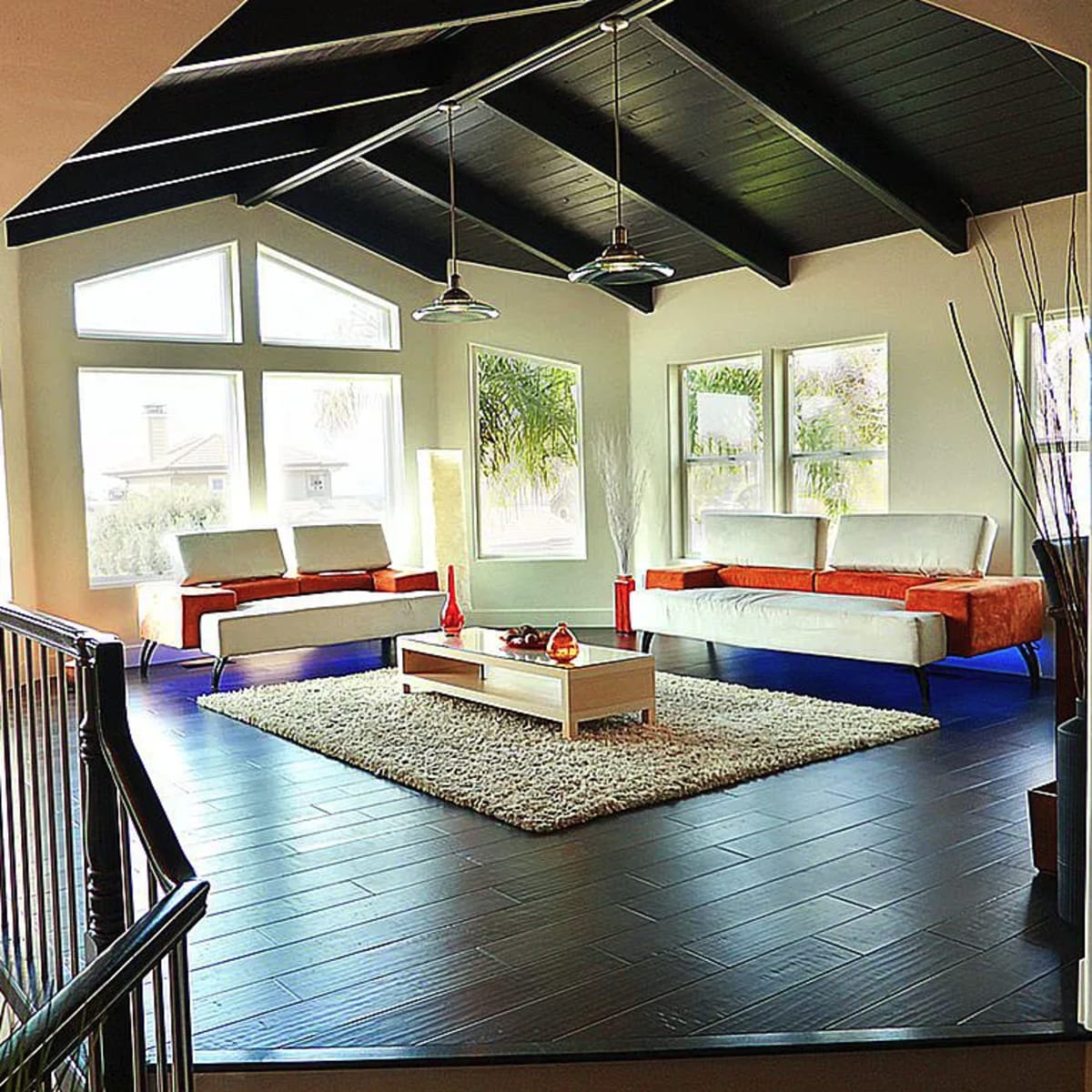A modern living room with a high ceiling, featuring two sofas, a coffee table on a rug, and large windows allowing natural light.