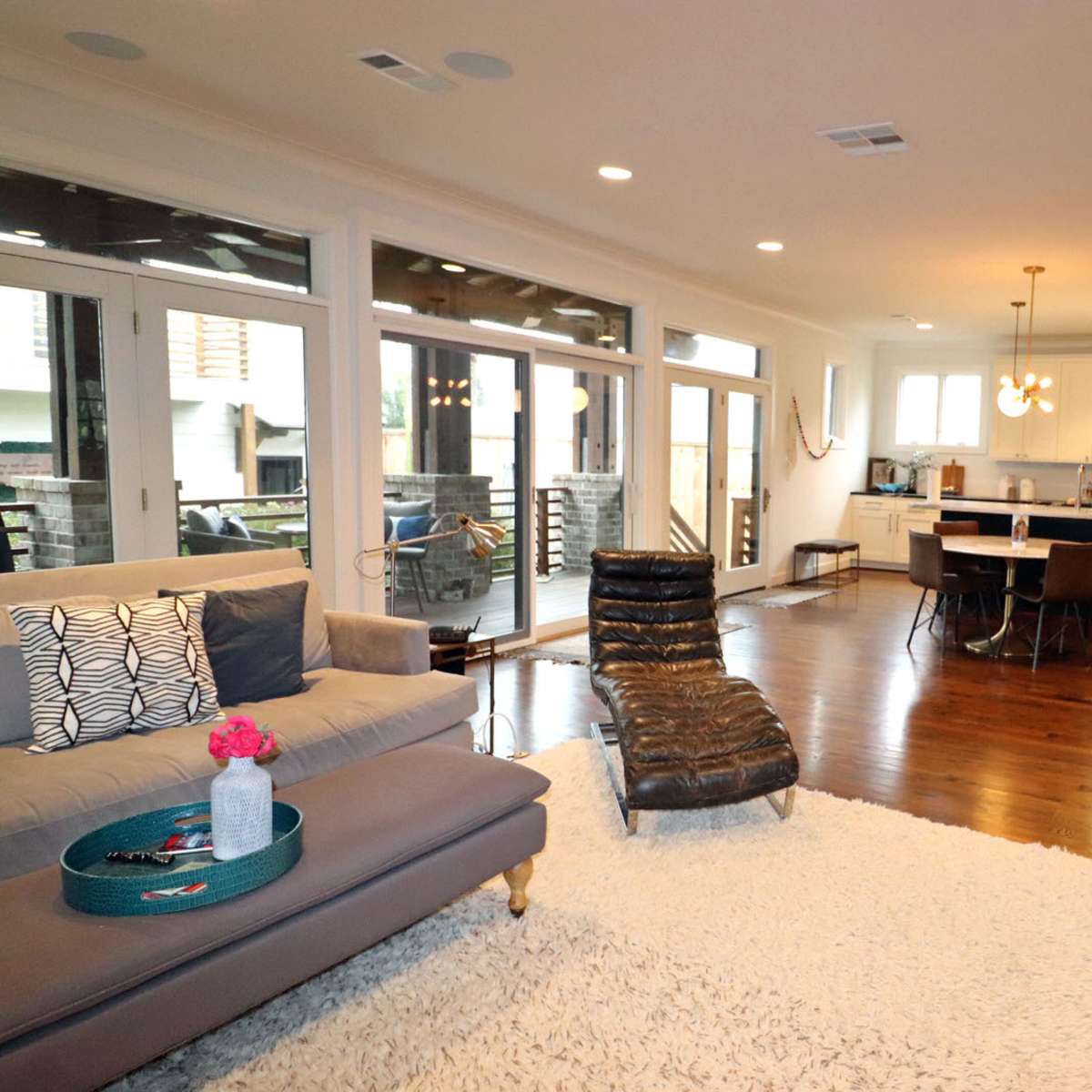 A modern living room and kitchen area featuring hardwood floors and large windows that illuminate the space.