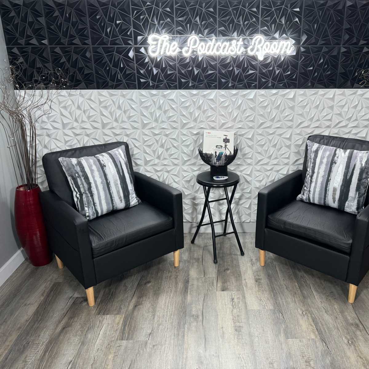 Two black armchairs with striped cushions and a small black table between them, set against a textured wall featuring the text "The Podcast Room" in neon lighting.