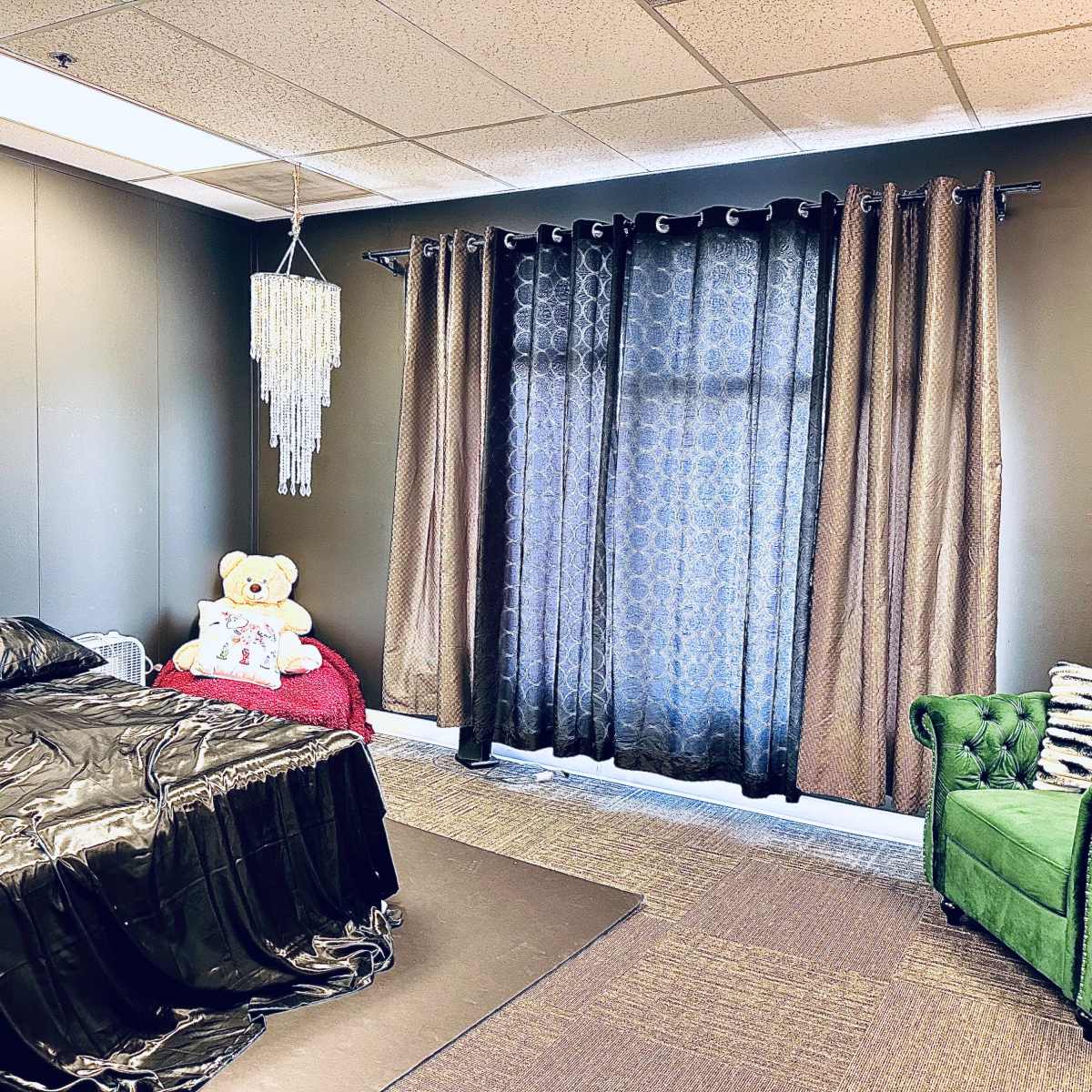 A bedroom featuring a black bedspread, a teddy bear on the bed, a green chair, and dark curtains covering a window.