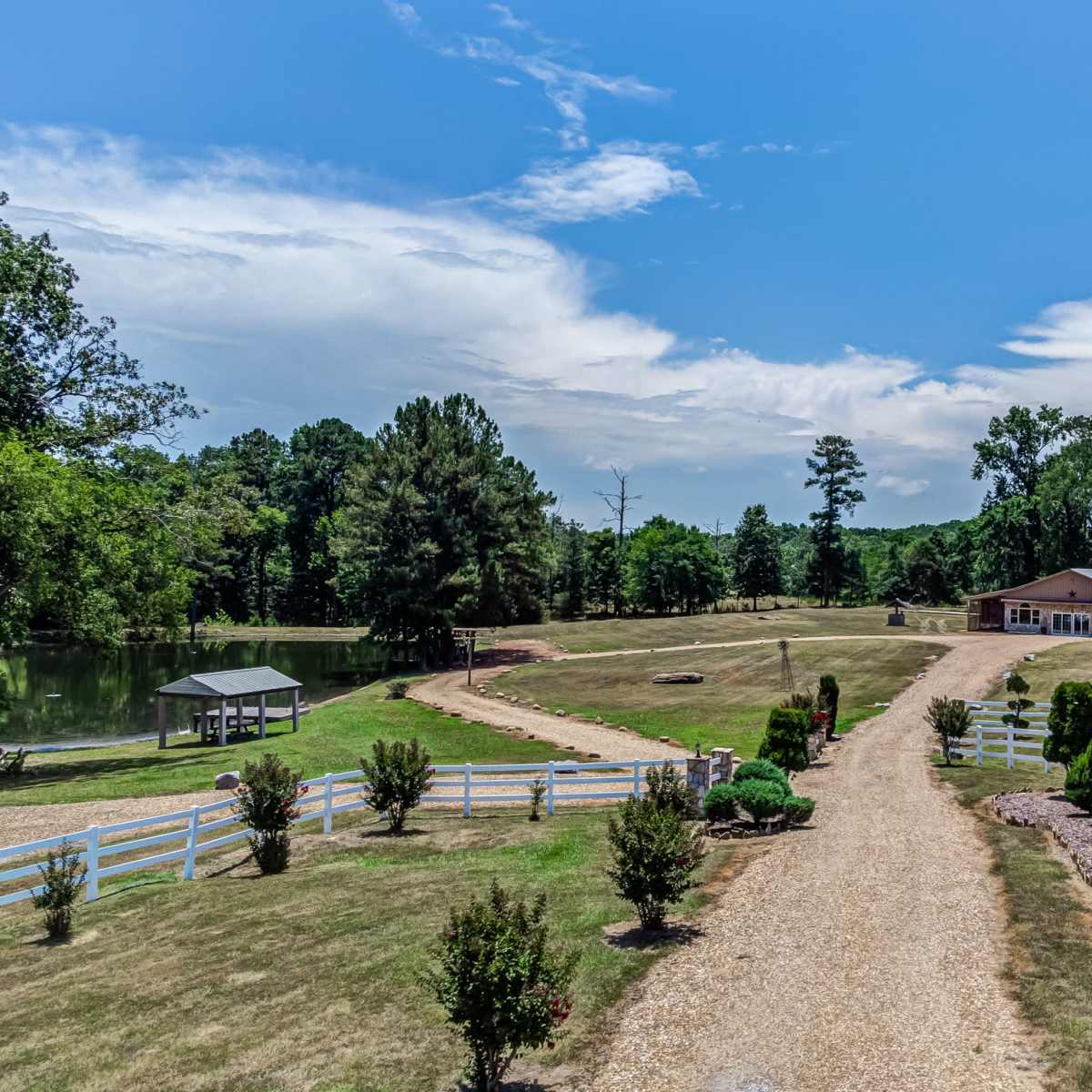 A spacious landscape with a gravel driveway leading to a large building, surrounded by manicured greenery and a pond on the left.