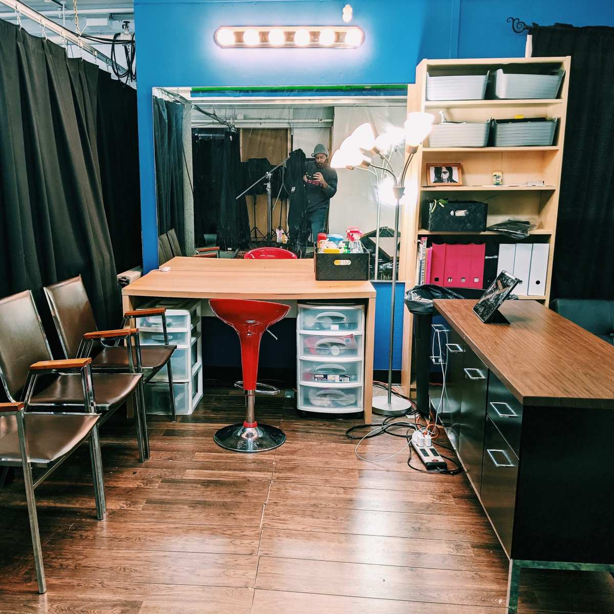 A small studio space with a wooden table, a red bar stool, several chairs, a desk with storage, and black curtains.