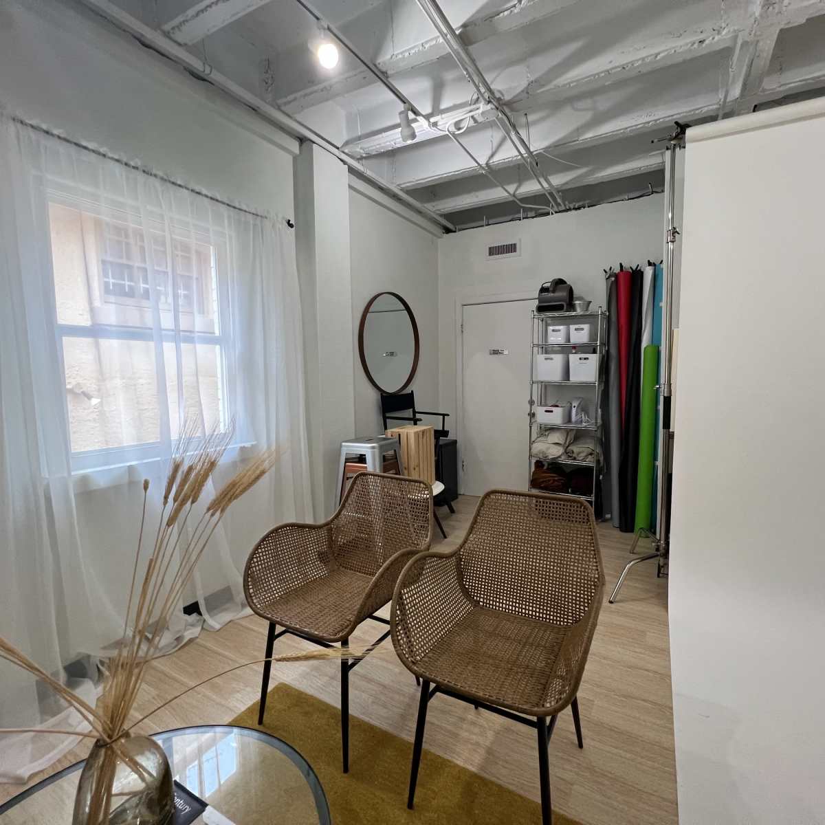 A minimalist room with two woven chairs facing a window, a round mirror on the wall, and storage containers in the corner.
