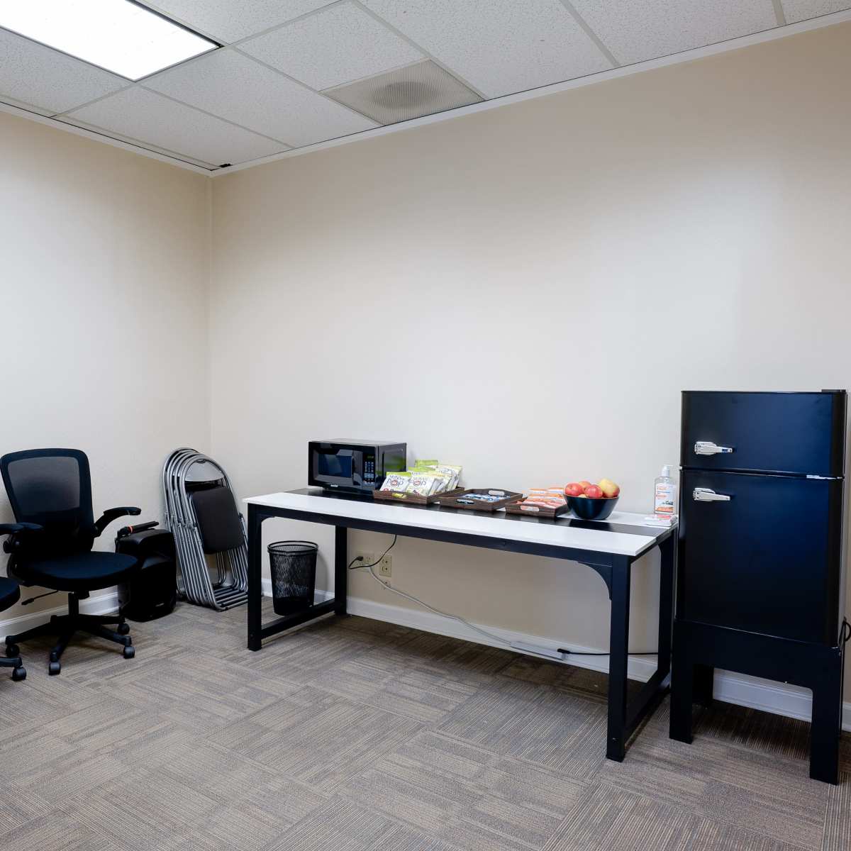 A small office space with two black chairs, a table displaying snacks and a microwave, and a black refrigerator along the wall.
