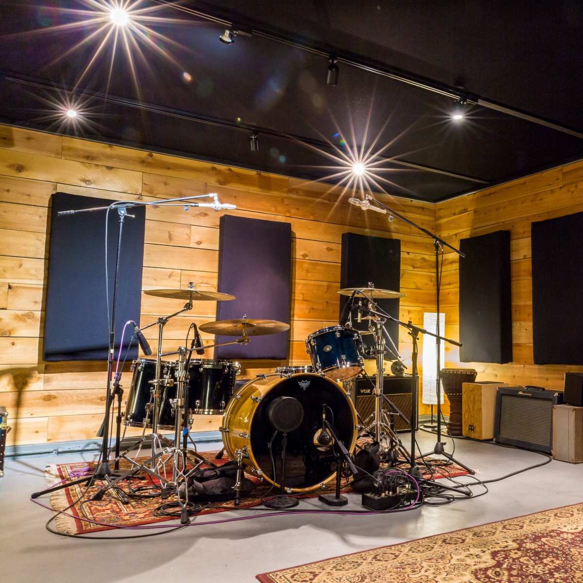 A recording studio features a drum set, microphones, and amplifiers against wooden paneling and soundproofing.