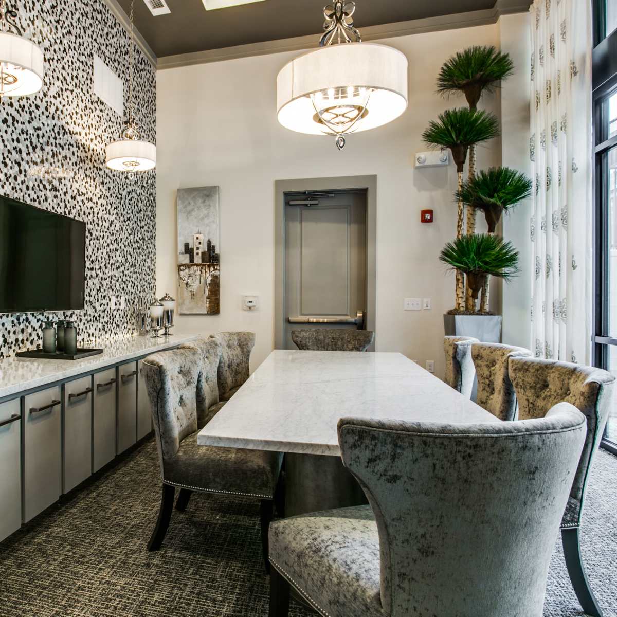 A modern dining area with a long marble table, plush chairs, a patterned accent wall, and a wall-mounted television.
