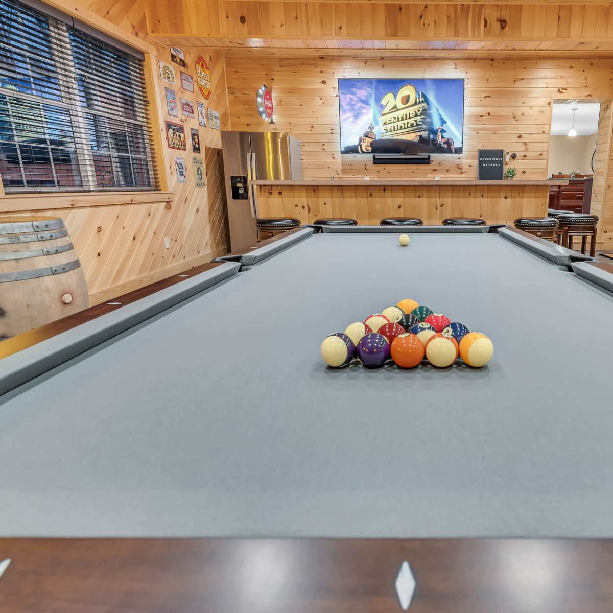 A billiards table with a tight triangle of colored balls is set in a wooden-paneled game room that features a television mounted on the wall.