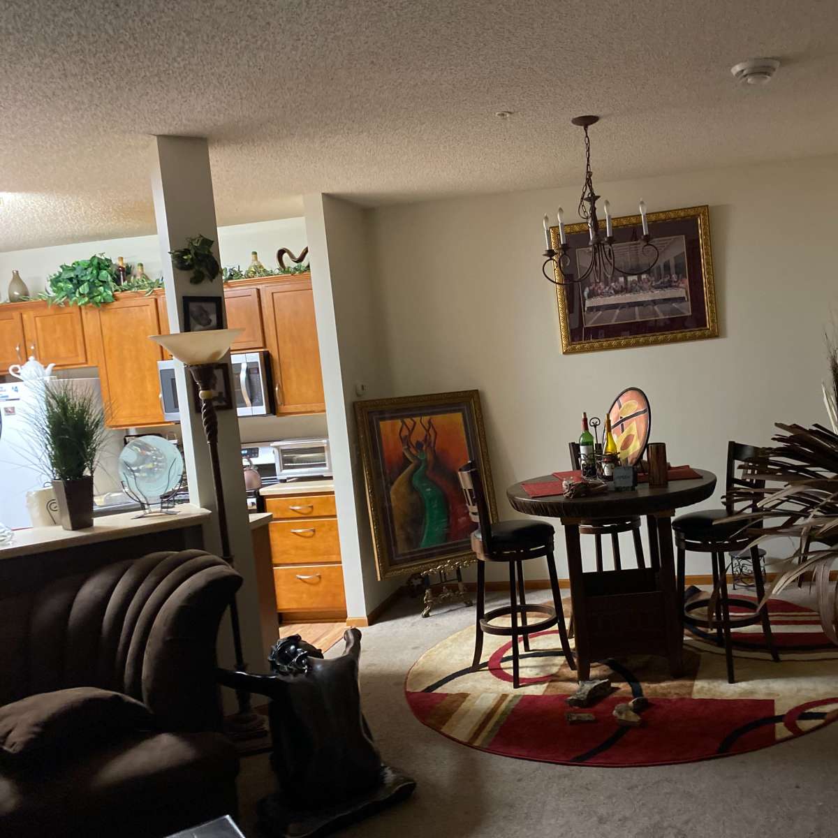 A living room and dining area with a round table and chairs, a large painting, and kitchen cabinets in the background.