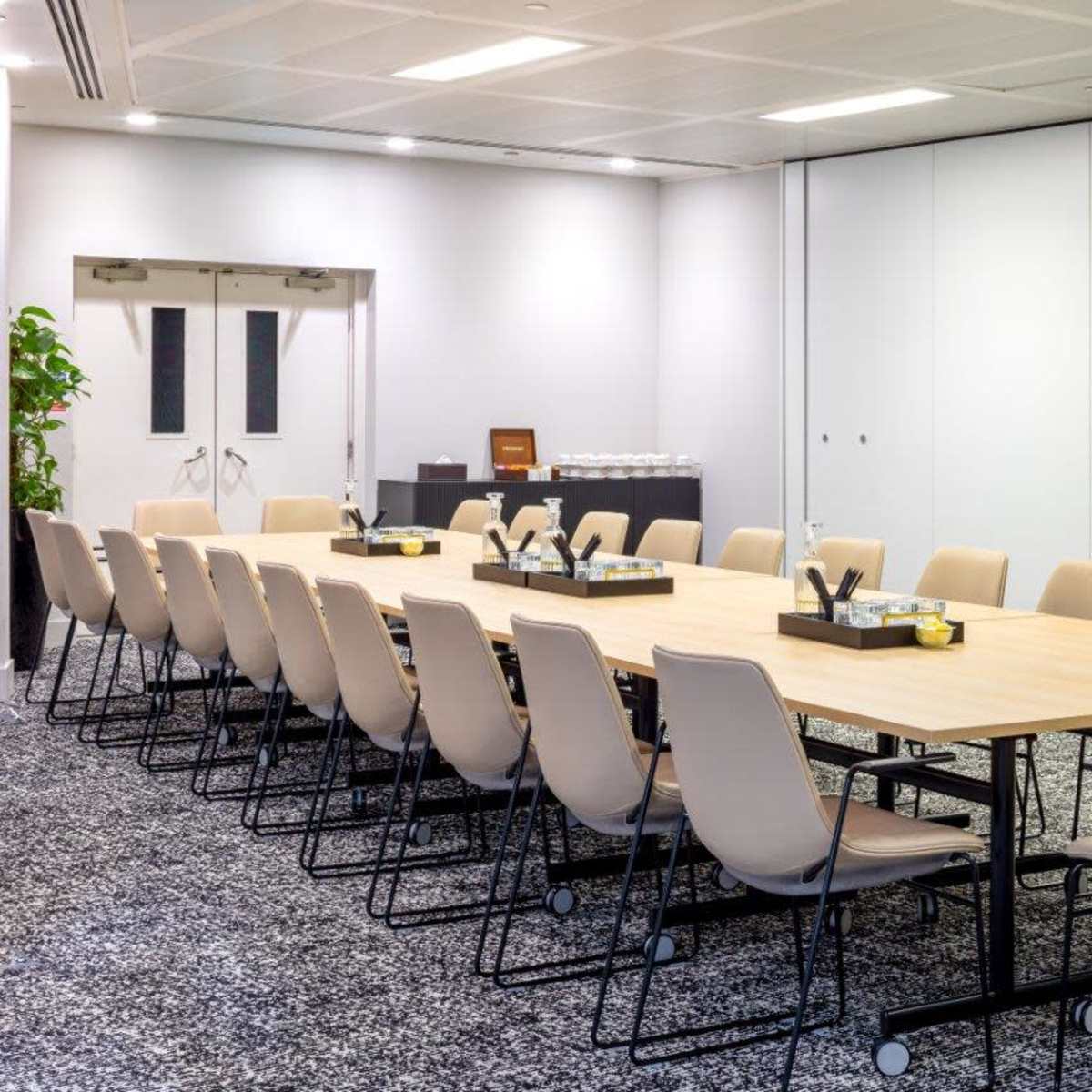 A large, rectangular conference table is set up in a meeting room with beige chairs arranged around it, alongside a refreshment station in the background.