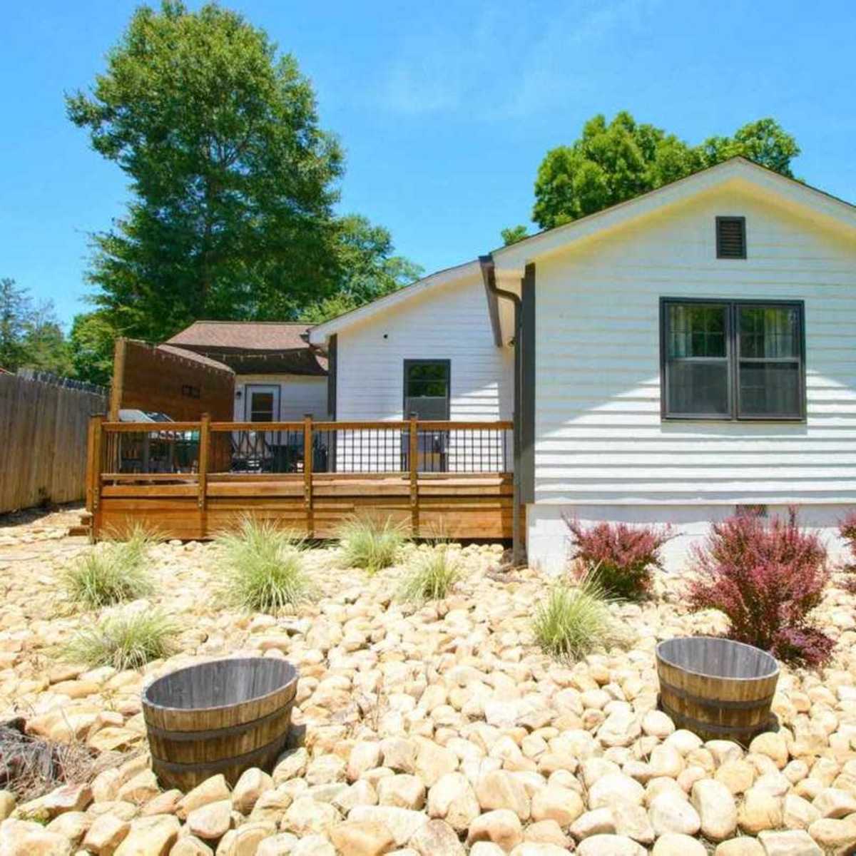 A single-story white house with a wooden deck surrounded by a landscaped yard featuring rocks, shrubs, and two wooden barrels.