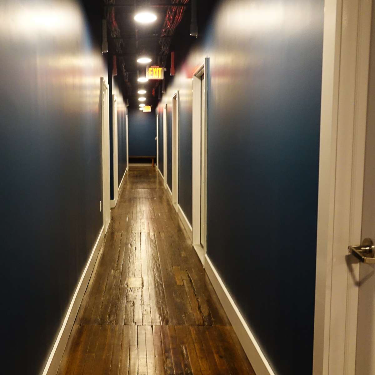 A narrow corridor with dark blue walls and wooden flooring, lined with doors on both sides and lit by overhead lights.