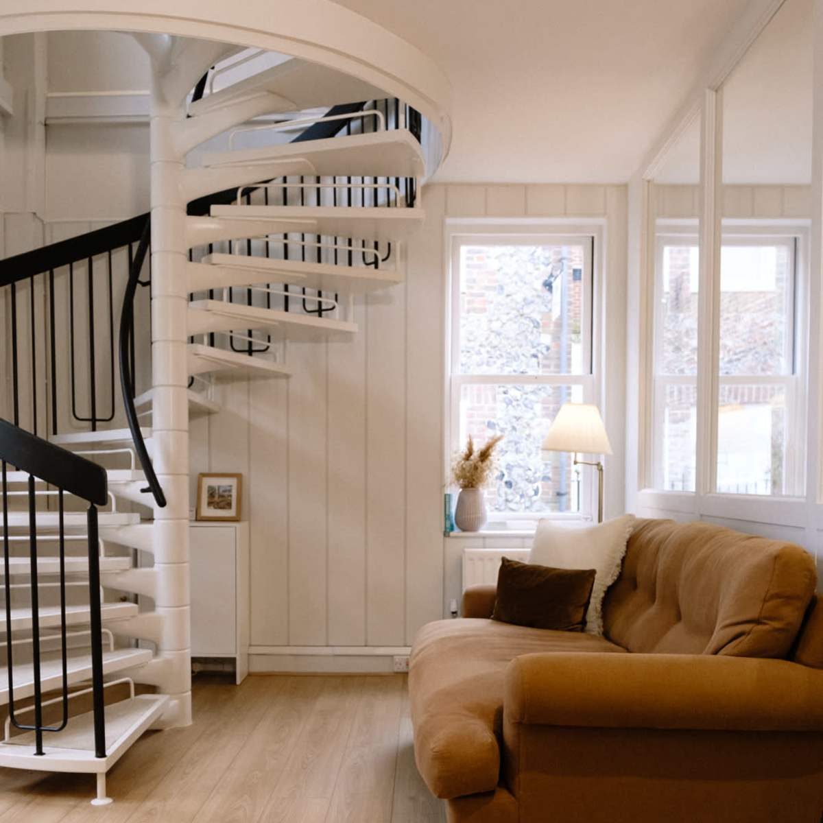 A spiral staircase leads from a light-colored room with wooden flooring to a cozy seating area featuring a brown couch and a lamp.