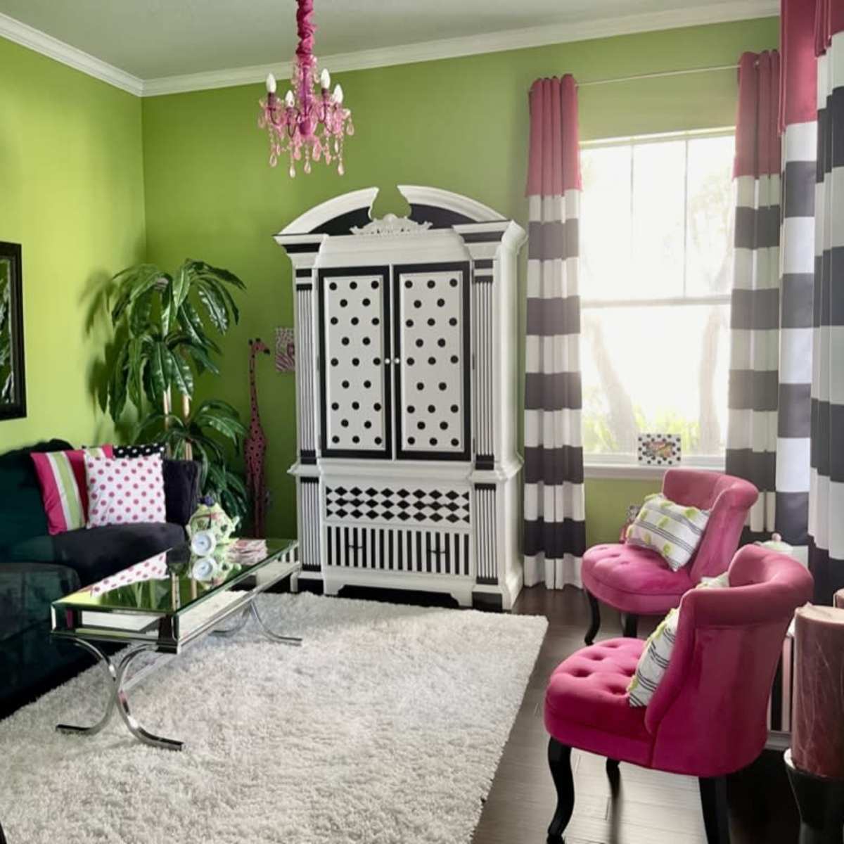 The room features green walls, a black velvet sofa with colorful pillows, a white cabinet with polka dots, and bright pink chairs, all set on a large white rug.