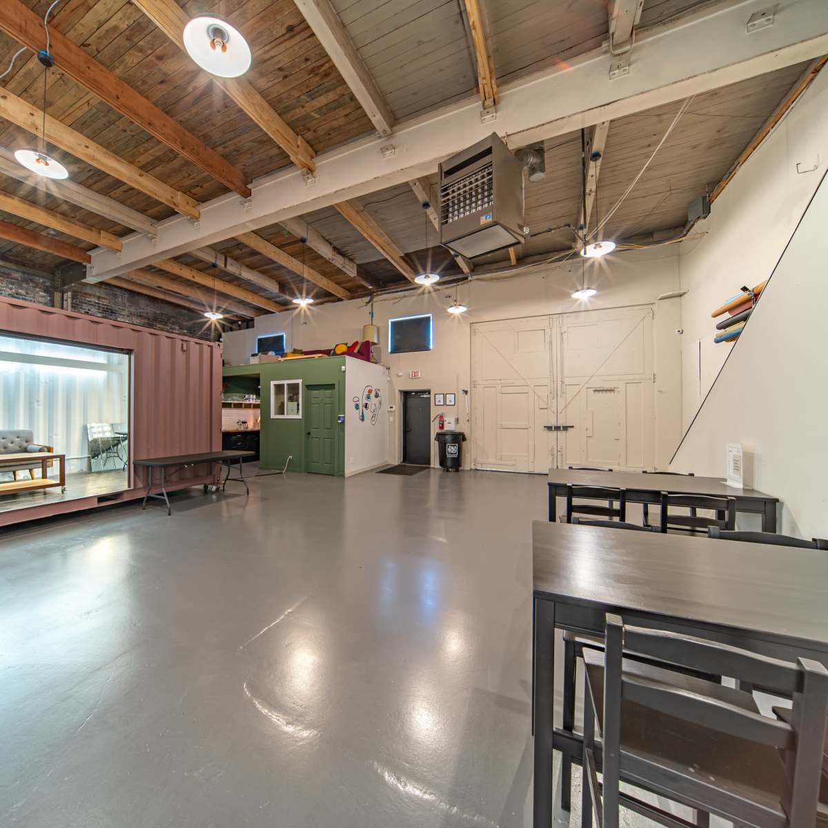 A spacious room with industrial-style wooden beams, featuring a shipping container, tables, and various furniture pieces.