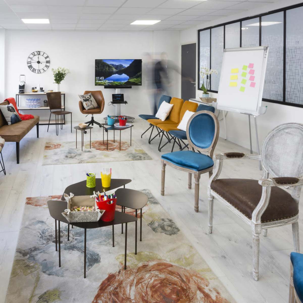 A modern, spacious meeting room filled with various seating options and tables, featuring large windows and a presentation area with a whiteboard.