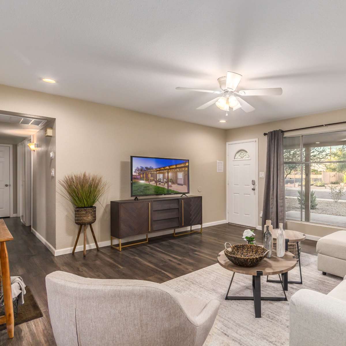 A modern living room features a white sectional sofa, a coffee table, and a television on a dark wood cabinet, with large windows allowing natural light.