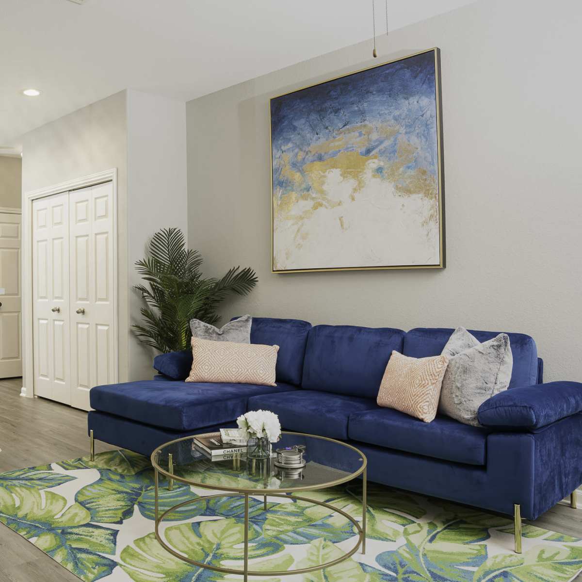A modern living space features a blue couch, a round glass coffee table, and a large painting on the wall, with an adjacent kitchen visible in the background.