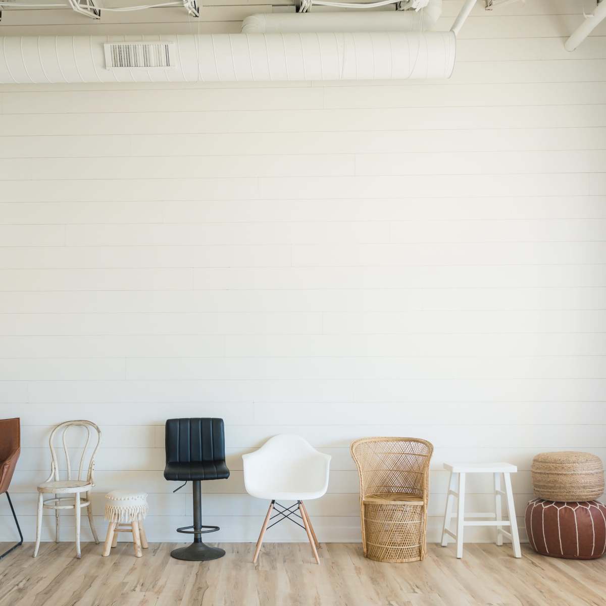 A row of five different styles of chairs and stools against a plain, white wall in a well-lit room.