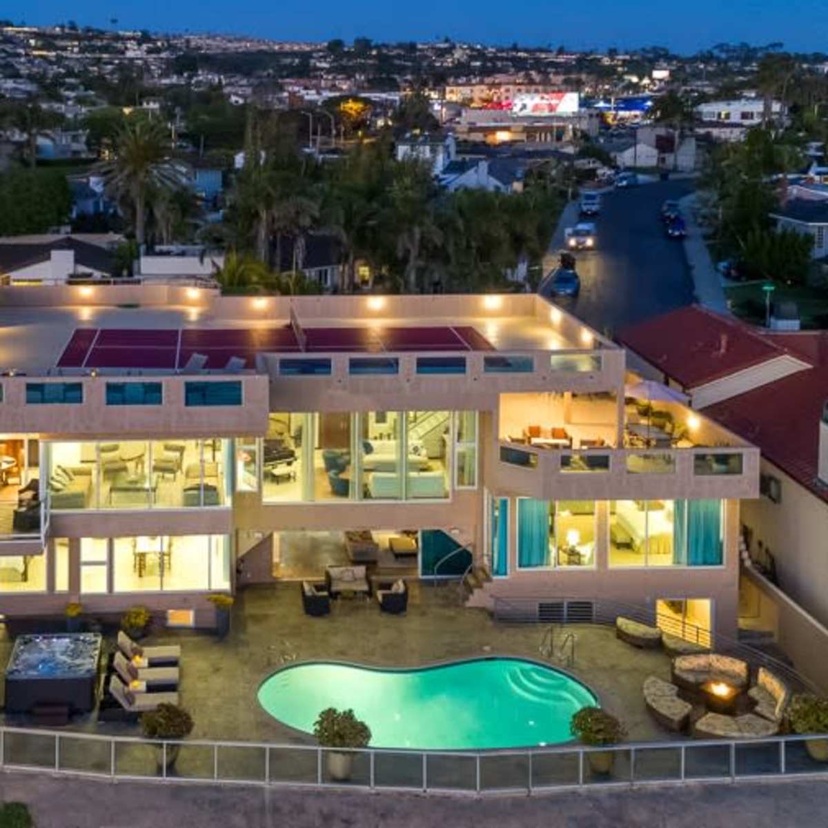 A modern multi-level house with large glass windows is situated beside a swimming pool, surrounded by palm trees and overlooking a suburban neighborhood at dusk.