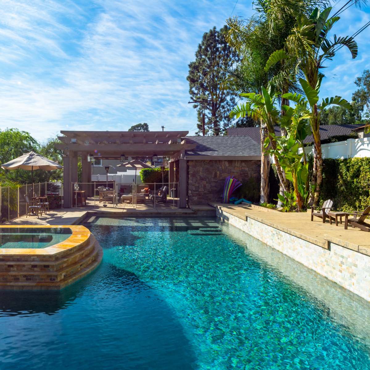 A residential backyard features a swimming pool with a spa, surrounded by lounge chairs and a pergola.