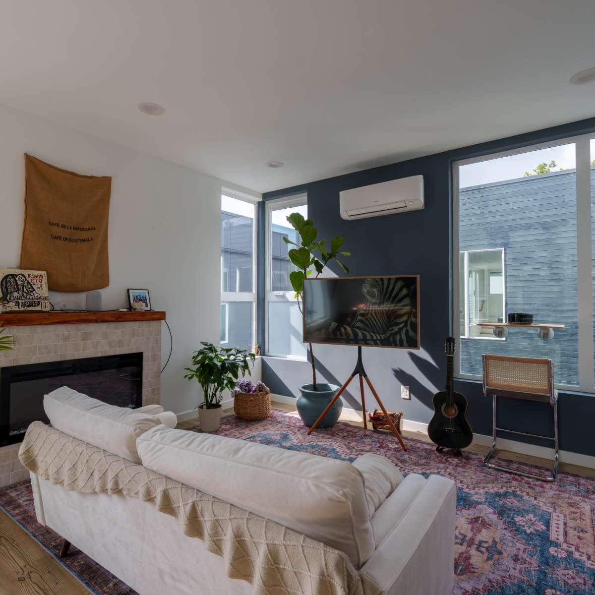A modern living room features a white couch, a fireplace, a large television mounted on a wall, and indoor plants, with large windows allowing natural light.