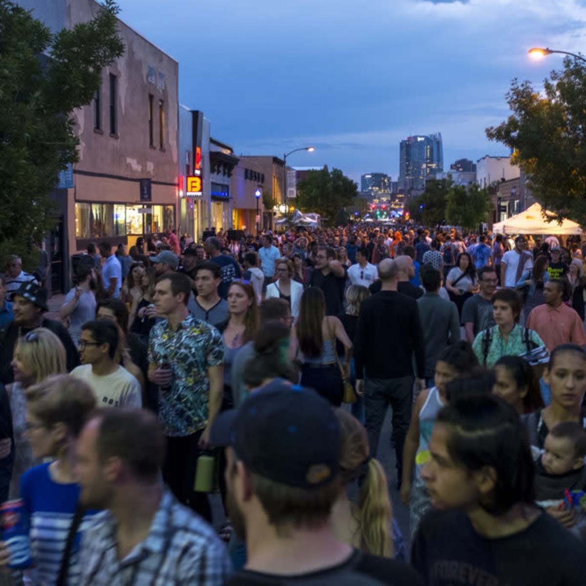 A large crowd of people fills a street with shops and buildings, under a dusk sky.
