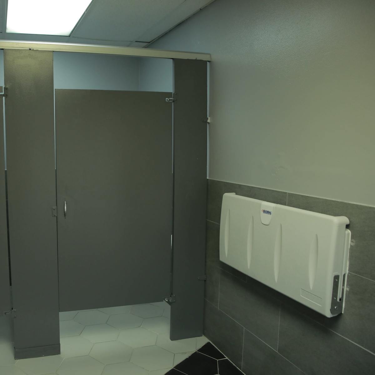 A bathroom stall area with three gray partitions, a mirror, a trash bin, and a changing table mounted on the wall.