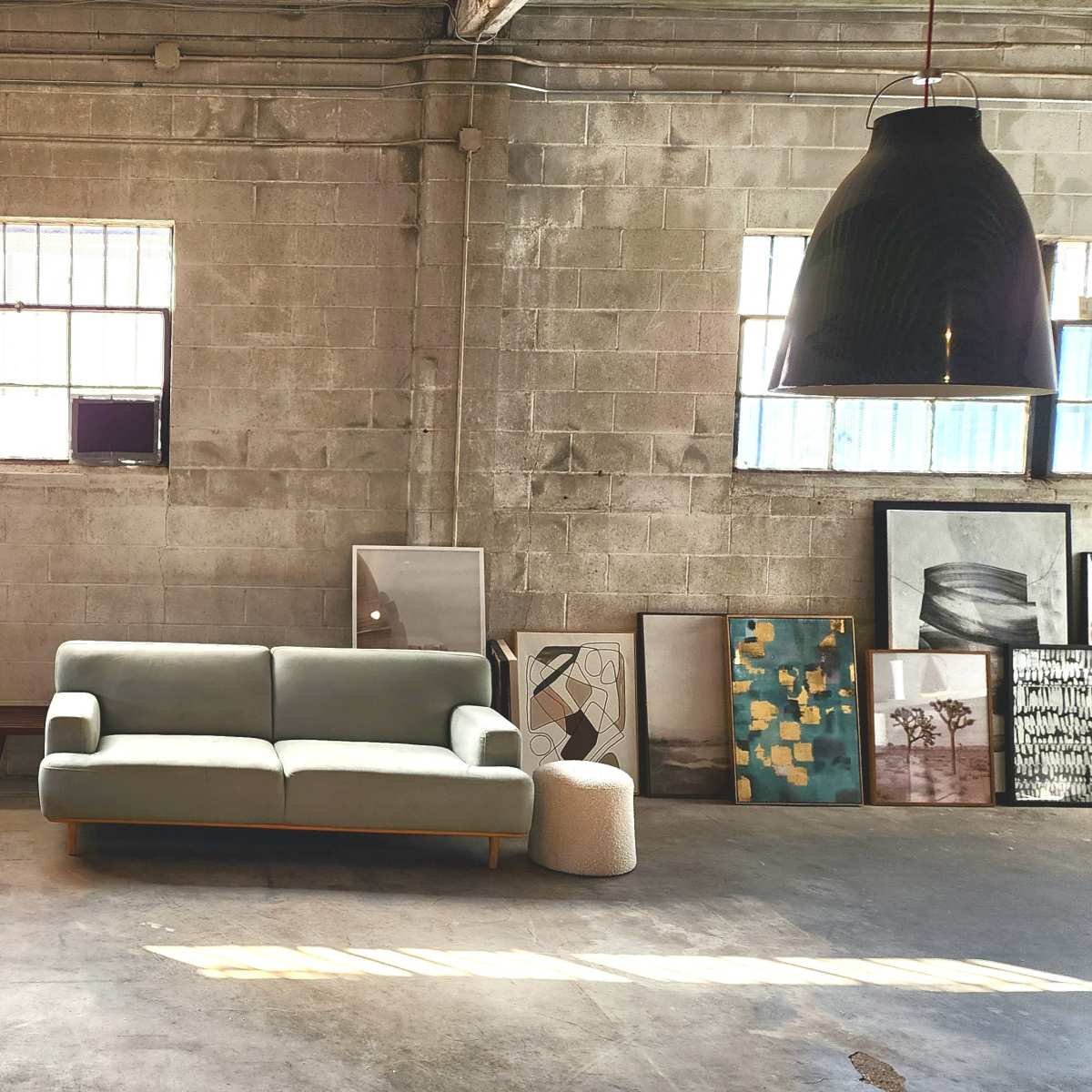 A light green sofa sits in the center of an industrial space surrounded by various framed artworks against the walls.
