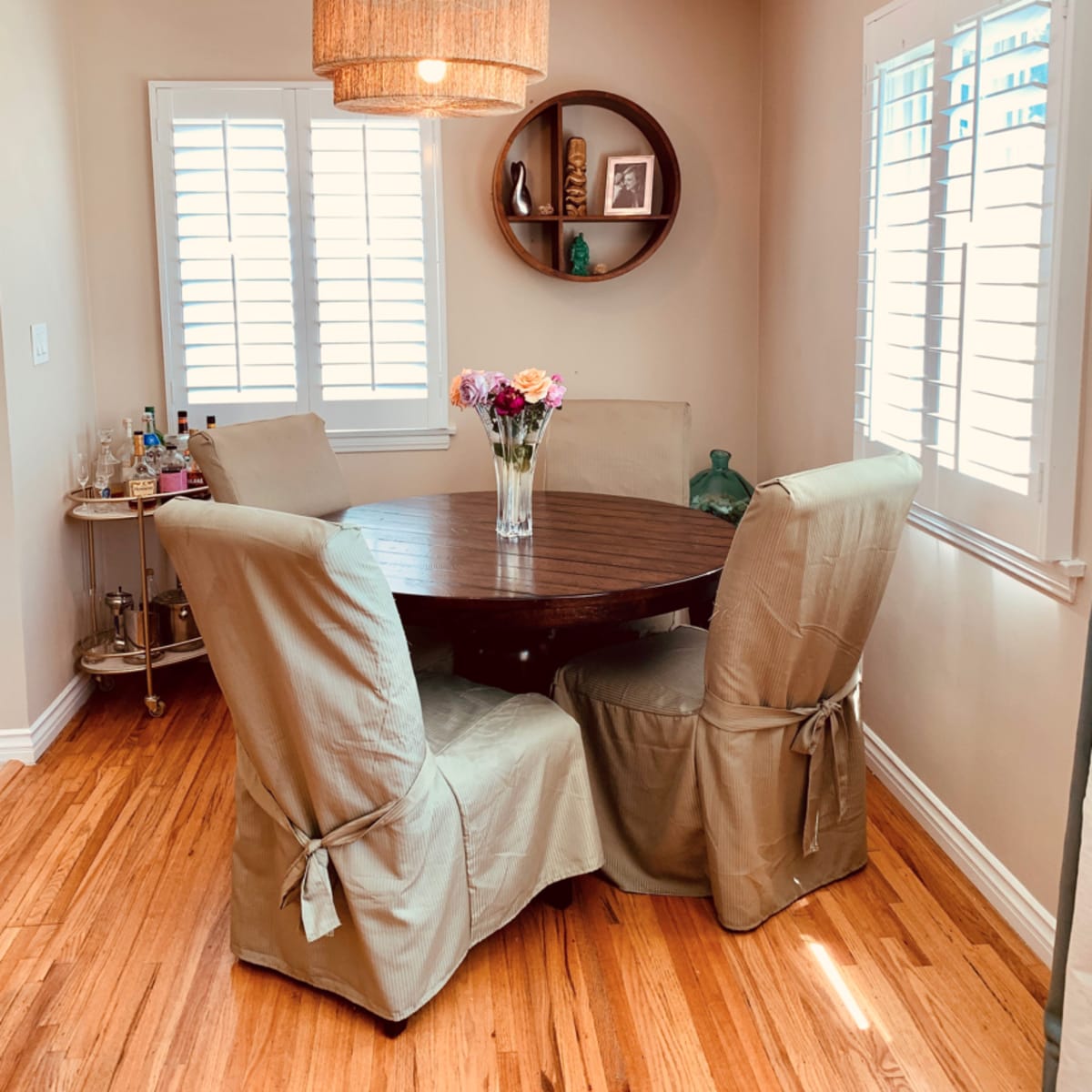 A round wooden dining table with four beige upholstered chairs is set in a bright room featuring a decorative light fixture and a wall shelf.