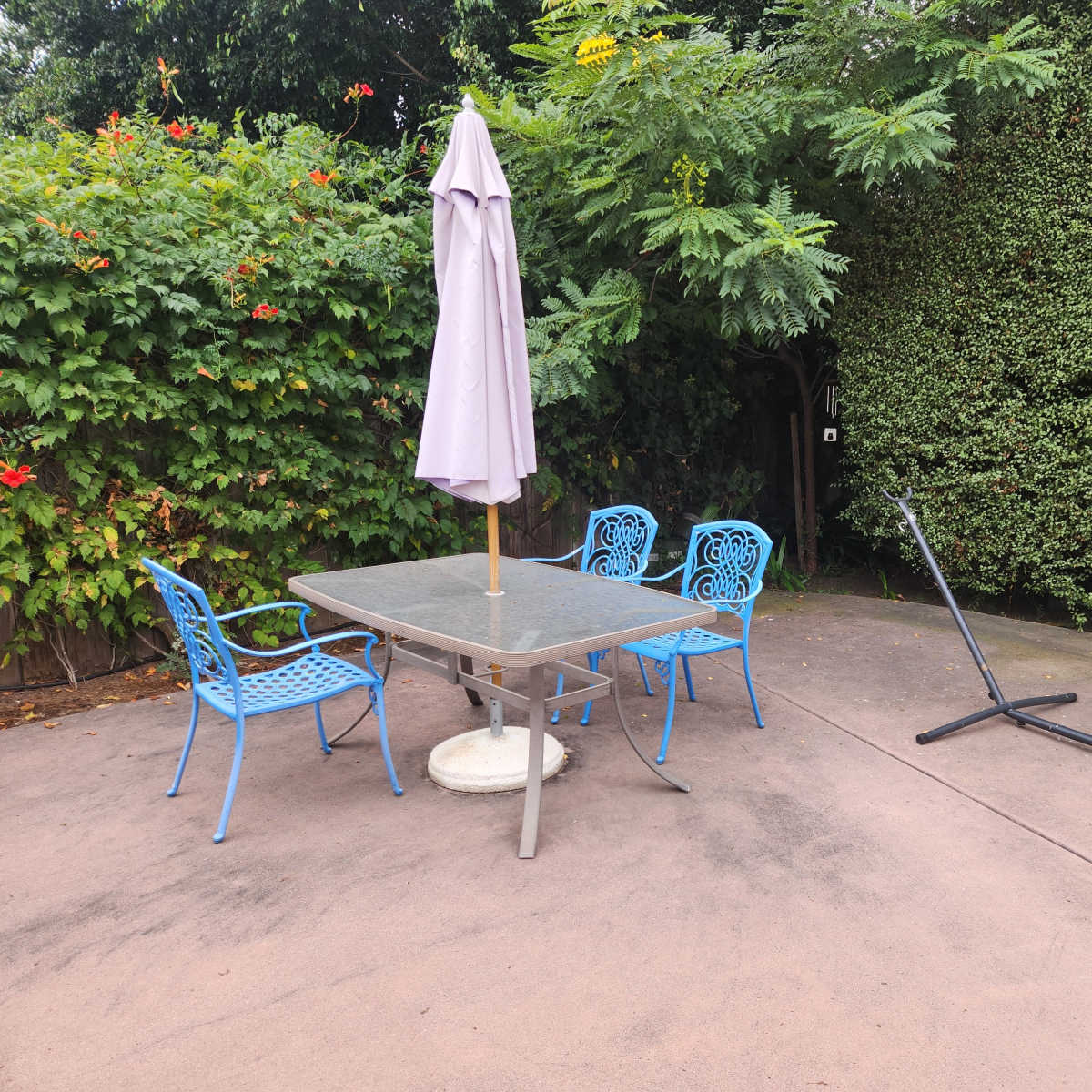 A patio area features a table with four blue chairs and an umbrella, alongside a disassembled hammock stand amidst greenery.