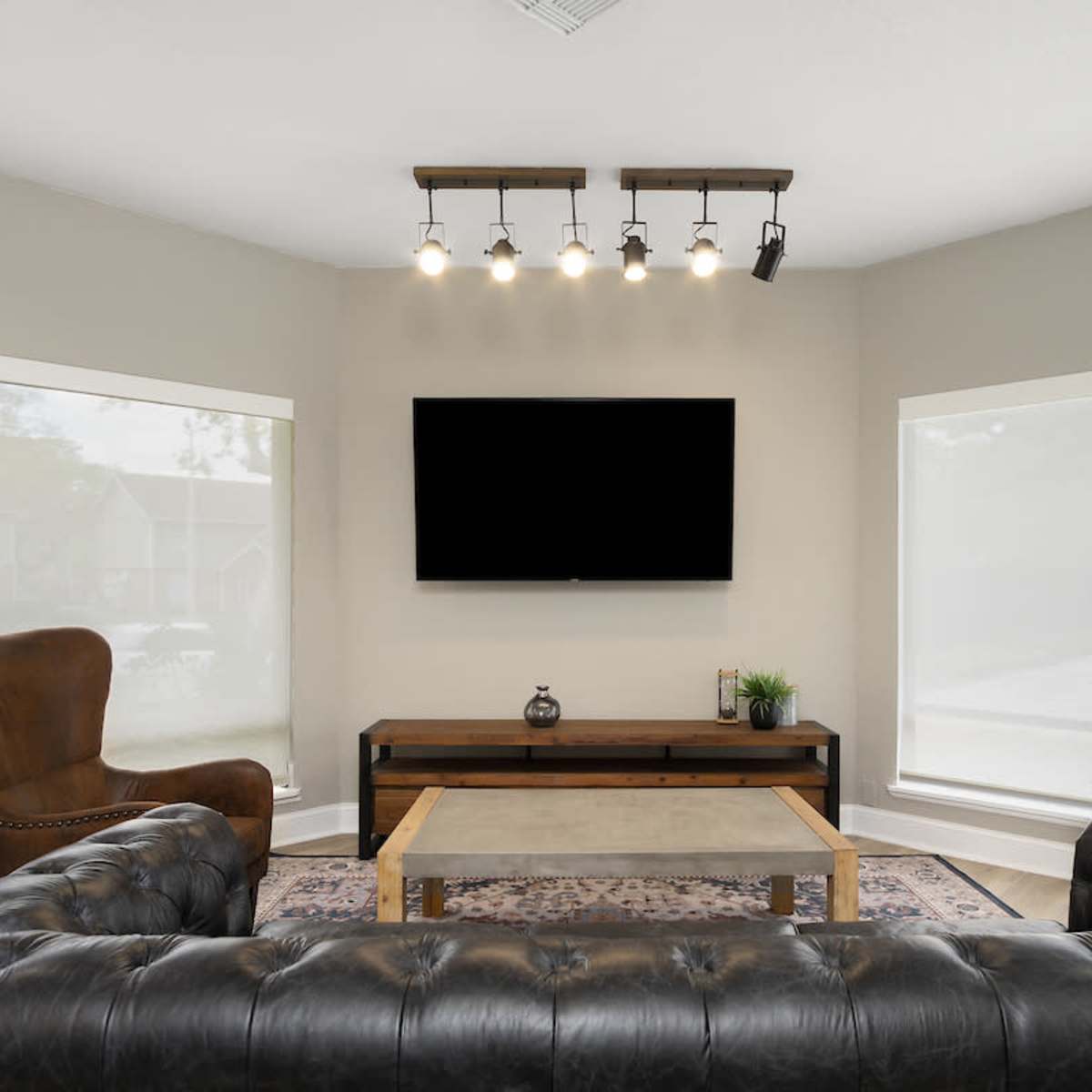 A modern living room features a leather sectional couch, two armchairs, a coffee table, and a wall-mounted television, illuminated by track lighting.