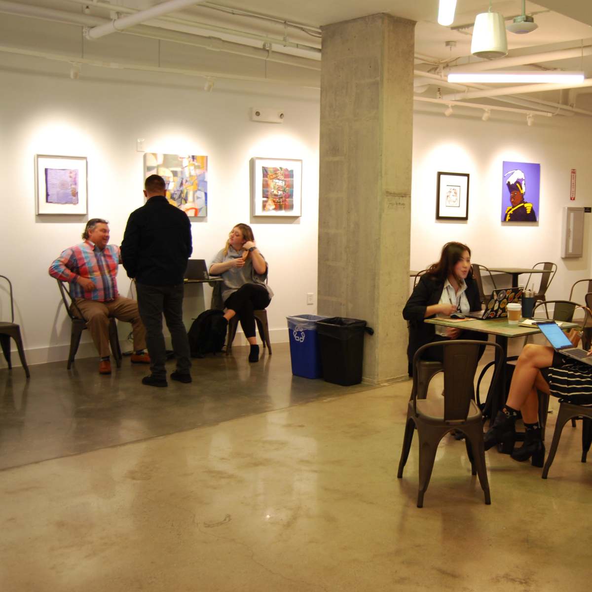 A modern workspace with people engaged in discussions and working on laptops at tables, with framed artworks on the walls.