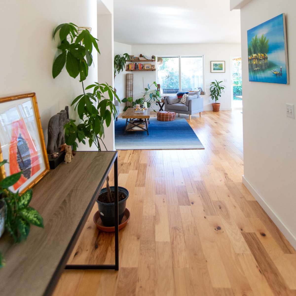A hallway features a wooden console table with decorative items, leading to a bright living area with plants and a view of a blue rug.
