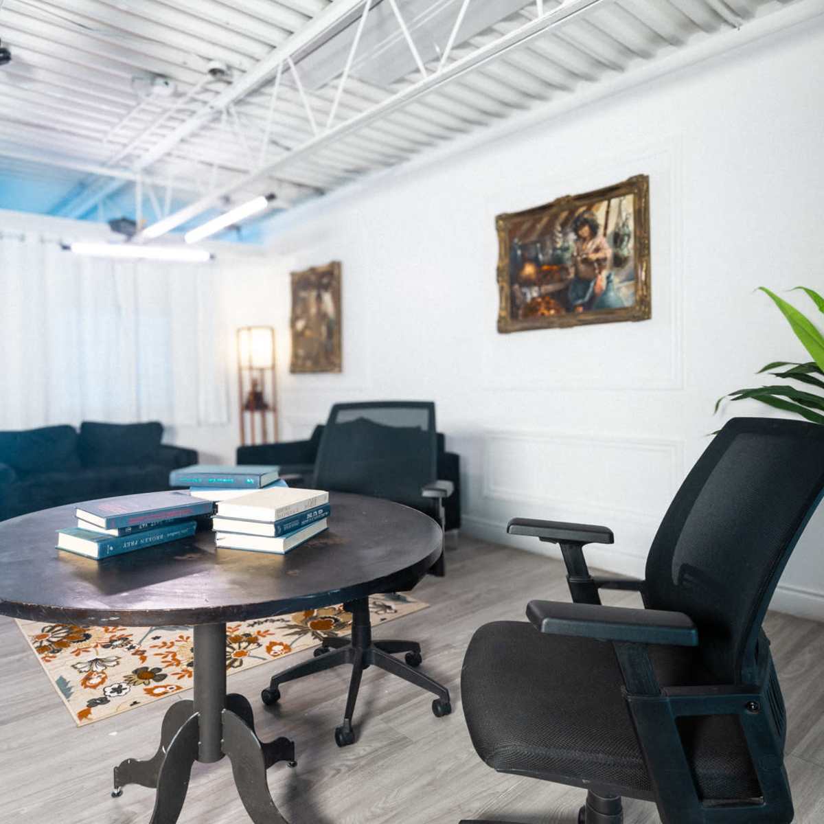 A spacious, well-lit office or lounge area with a round table, two black chairs, a potted plant, and a large screen in the background.