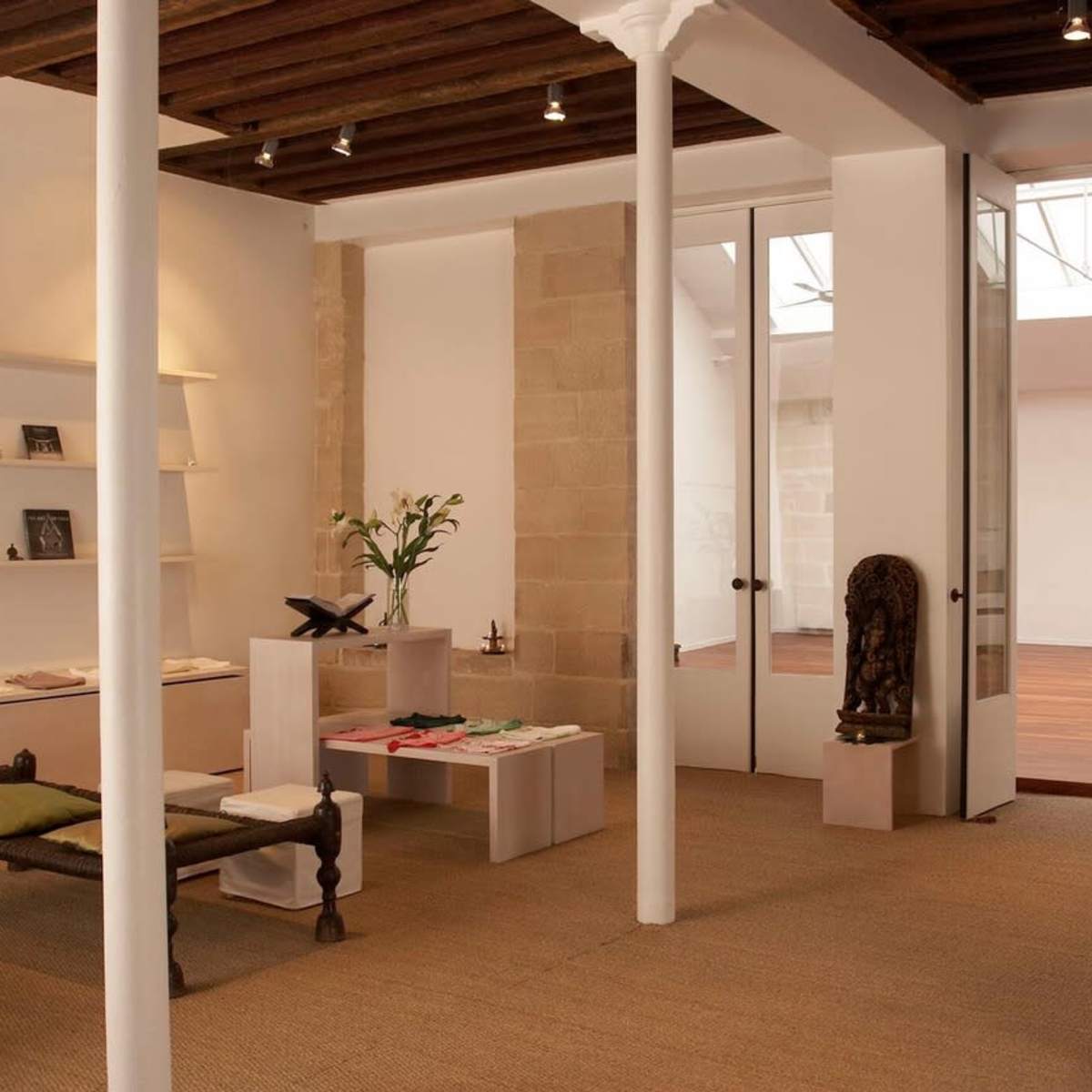 A minimalist interior of a gallery or boutique space with natural light, wooden beams, and shelves displaying various items.