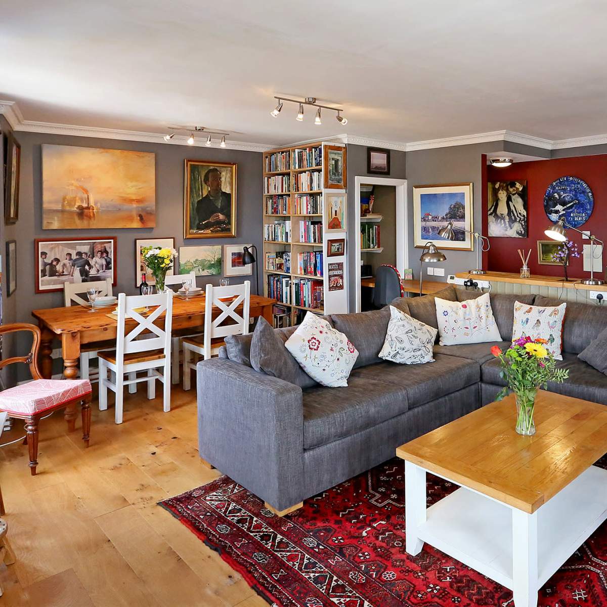 The scene shows a cozy living room with a gray sectional sofa, a wooden dining table, bookshelves filled with books, and various artworks on the walls.
