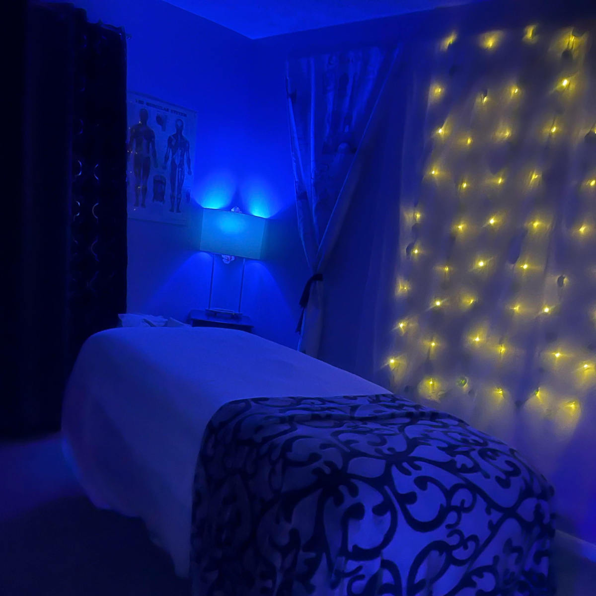 A bed with a dark patterned blanket is illuminated by blue light and surrounded by fairy lights in a dimly lit room.