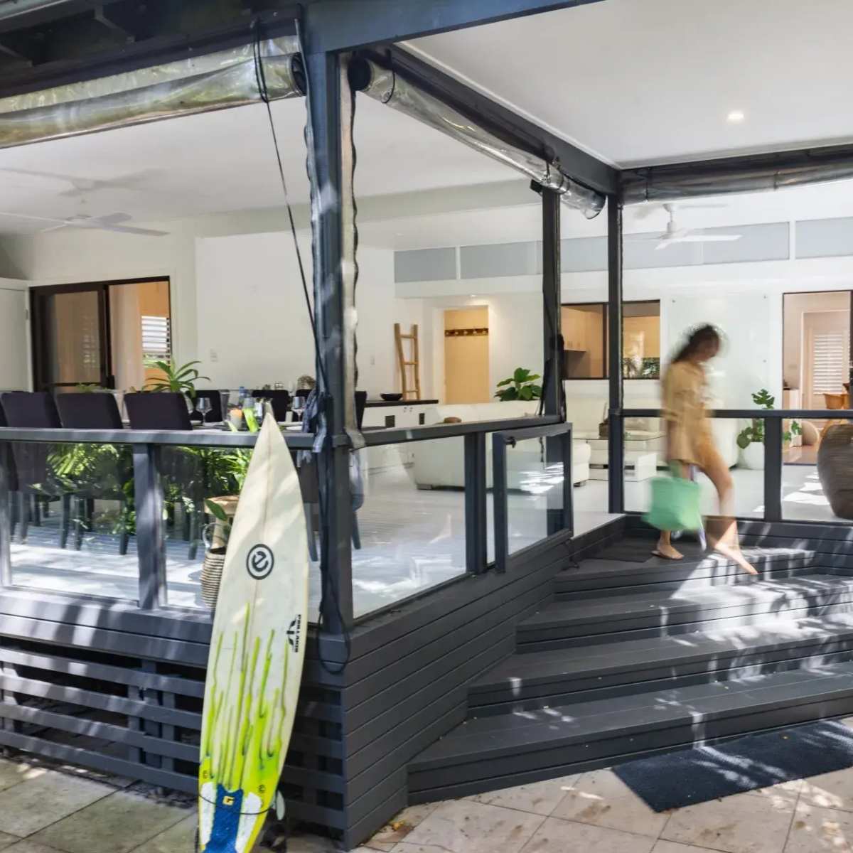 A person carries a bag while walking past a modern outdoor deck area of a house with sliding glass doors and potted plants.