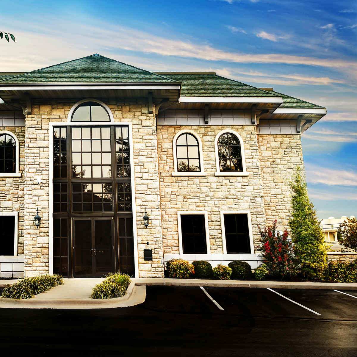 A large stone building with arched windows and a triangular roof is situated in a landscaped parking lot.